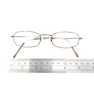 Puede incluir: Un par de gafas con una montura fina de metal oscuro y lentes transparentes. Las gafas est&aacute;n sobre una superficie blanca con una regla que muestra las medidas en cent&iacute;metros. La montura tiene forma rectangular.