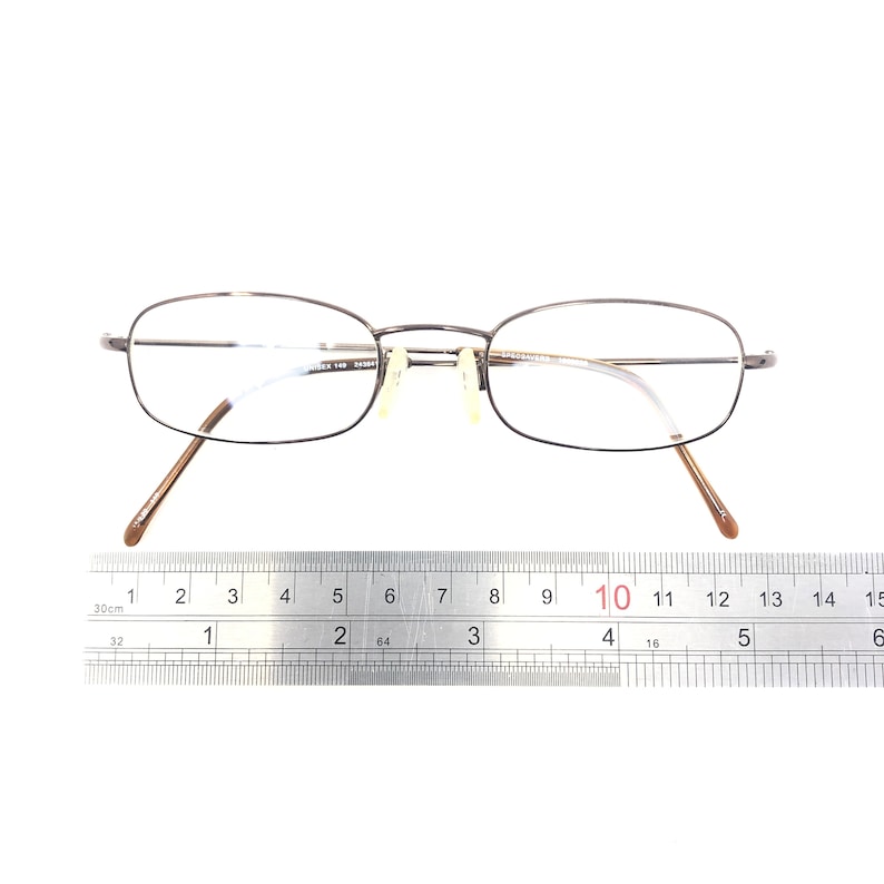 Puede incluir: Un par de gafas con una montura fina de metal oscuro y lentes transparentes. Las gafas est&aacute;n sobre una superficie blanca con una regla que muestra las medidas en cent&iacute;metros. La montura tiene forma rectangular.