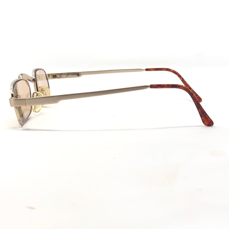 May include: A pair of eyeglasses with a rectangular, gold-colored frame and tinted lenses. The temples are a silver color, transitioning to a tortoiseshell pattern at the ear pieces. The glasses are set against a white background.