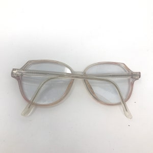 May include: Clear, vintage eyeglasses with a light pink tint around the lenses. The frames are a translucent, light gray color. The glasses have a classic, retro design.
