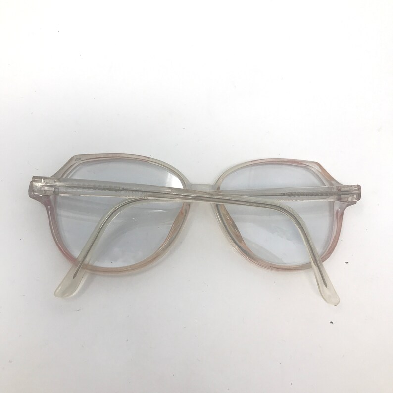 May include: Clear, vintage eyeglasses with a light pink tint around the lenses. The frames are a translucent, light gray color. The glasses have a classic, retro design.