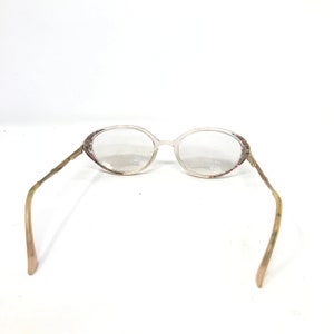 May include: Oval-shaped eyeglasses with a clear frame and lenses. The temples are a light brown color with a decorative pattern. The glasses are set against a white background.