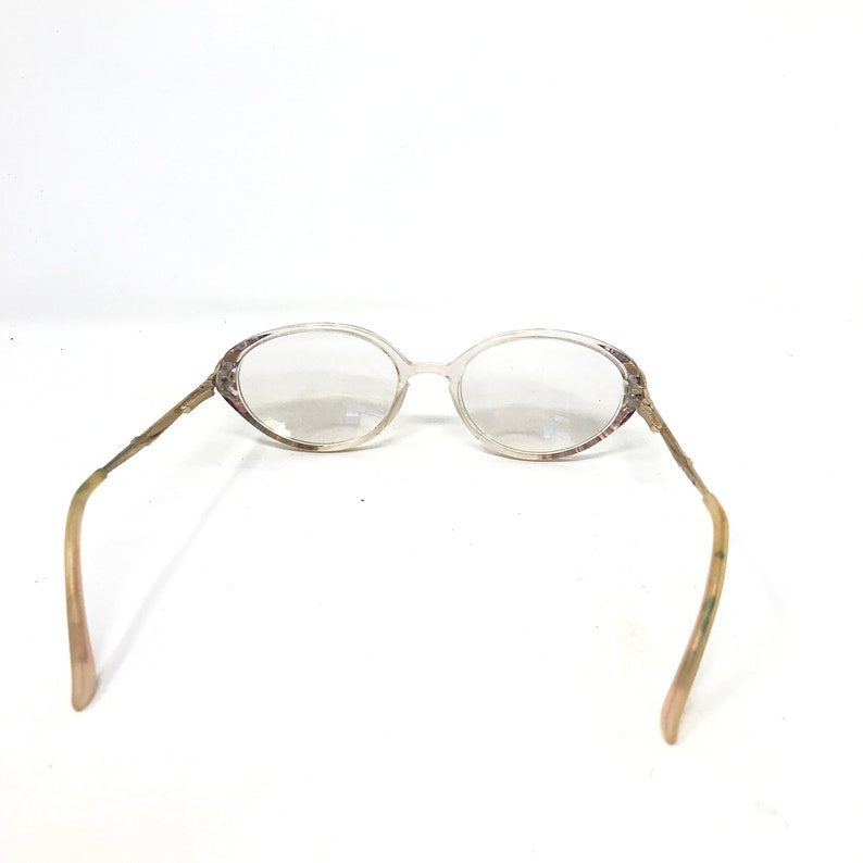 May include: Oval-shaped eyeglasses with a clear frame and lenses. The temples are a light brown color with a decorative pattern. The glasses are set against a white background.