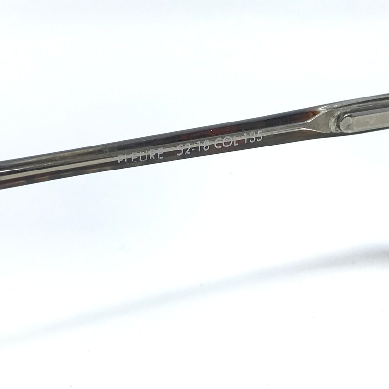 May include: Close-up of a silver-toned eyeglass frame. The text "PICTURE 52-18 COL 135" is etched on the temple arm. The frame is set against a white background.
