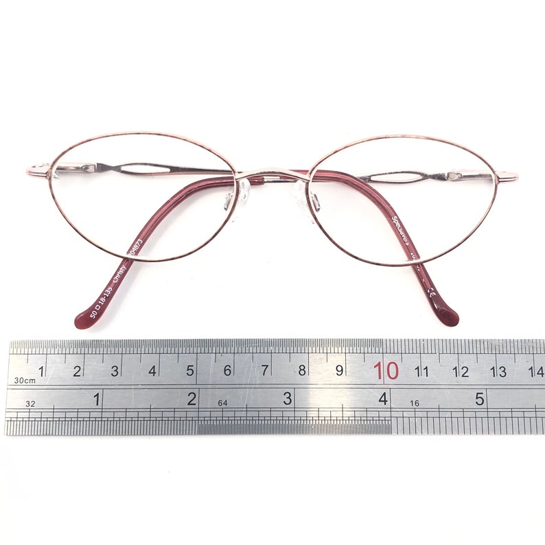 Puede incluir: Gafas ovaladas con una fina montura de metal marr&oacute;n rojizo. Las patillas y el puente nasal son de color marr&oacute;n rojizo a juego. Las gafas est&aacute;n sobre una superficie blanca con una regla.