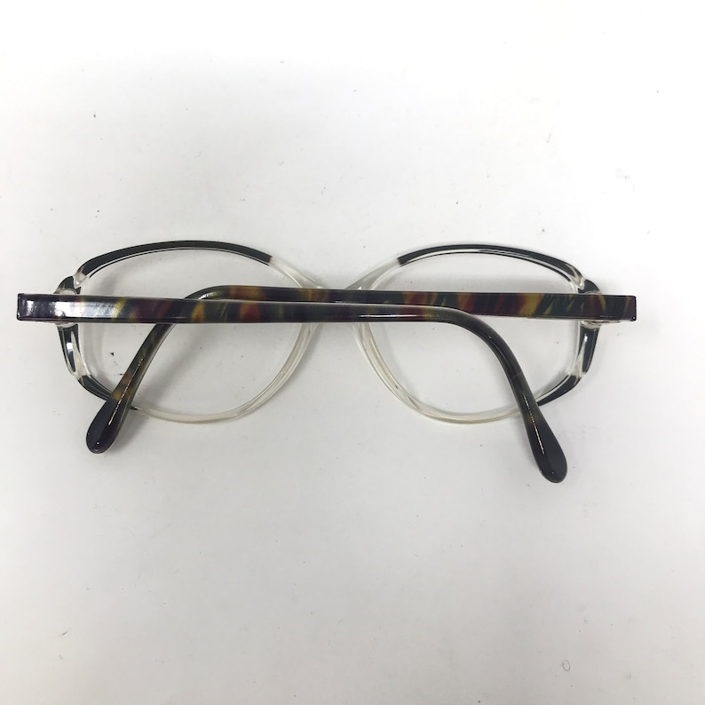 May include: Eyeglasses with a clear frame and a patterned brown, green, and yellow top bar. The temples are dark brown and the lenses are clear. The glasses are set against a white background.