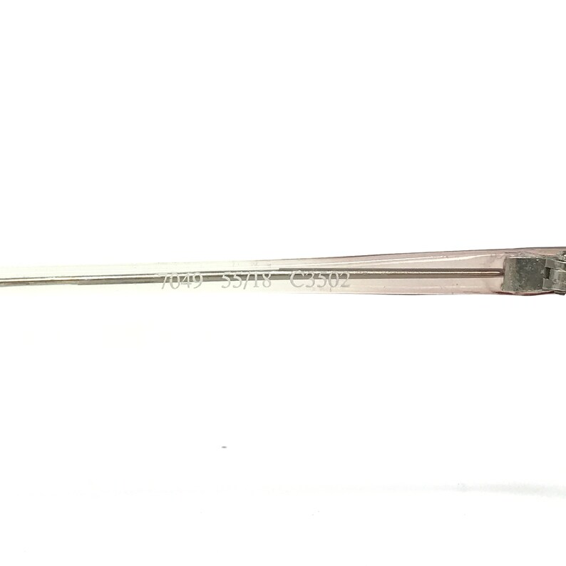 May include: Close-up of a transparent eyeglasses frame with the text "7049 55/18 C3502" printed on the arm. The frame is a light, neutral color, and the image is set against a white background.