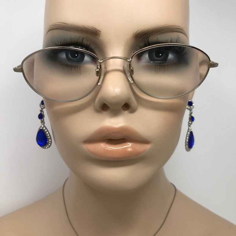 Puede incluir: Gafas ovaladas de color plateado con lentes transparentes. El maniqu&iacute; tambi&eacute;n lleva pendientes largos y colgantes con gemas azules en forma de l&aacute;grima y pedrer&iacute;a transparente. El tono de piel del maniqu&iacute; es claro y el fondo es blanco.