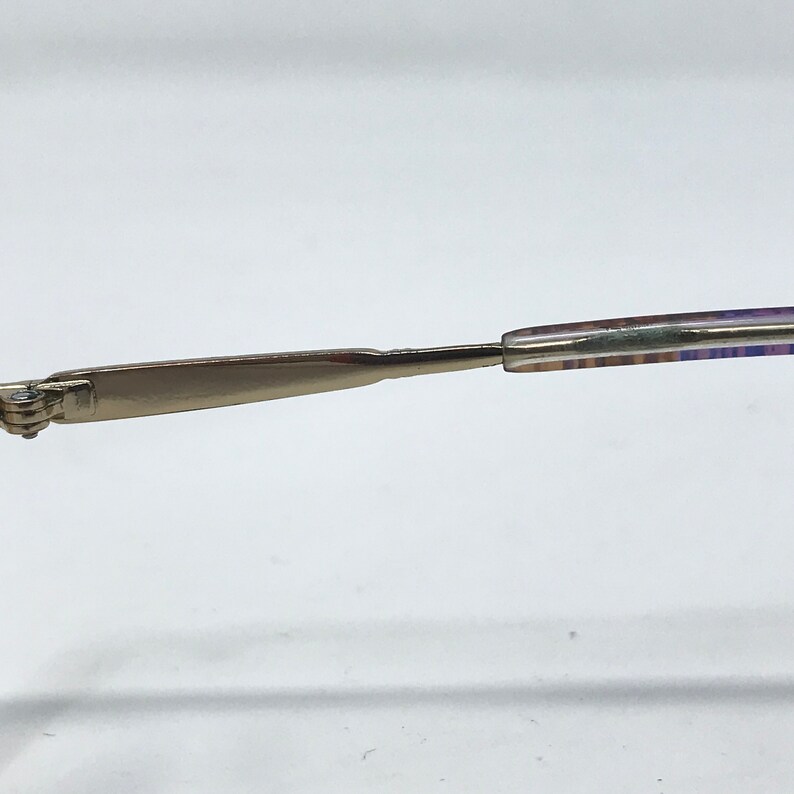 May include: Close-up of a silver-toned eyeglass frame arm. The arm has a flat, rectangular shape near the hinge and tapers to a thinner, patterned section. The patterned section has a mix of colors, including purple and blue.