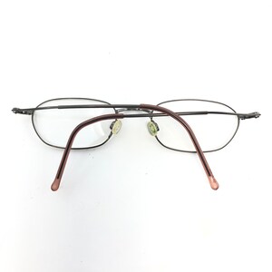 Puede incluir: Gafas ovaladas con montura de metal oscuro y lentes transparentes. Las patillas son de color marr&oacute;n rojizo. Las almohadillas nasales son de color amarillo claro. Las gafas est&aacute;n sobre un fondo blanco.
