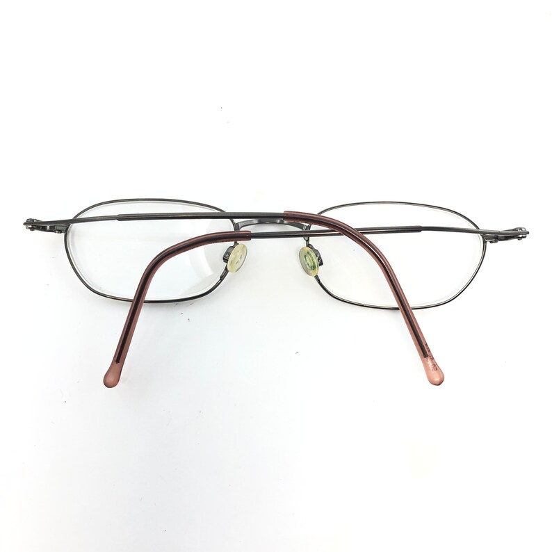 Puede incluir: Gafas ovaladas con montura de metal oscuro y lentes transparentes. Las patillas son de color marr&oacute;n rojizo. Las almohadillas nasales son de color amarillo claro. Las gafas est&aacute;n sobre un fondo blanco.