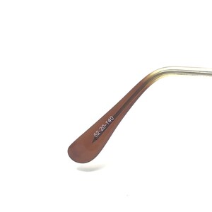 May include: Close-up of the arm of a pair of eyeglasses. The arm is a light brown color with the text "52-20-140" printed on it. The frame is a gold color.