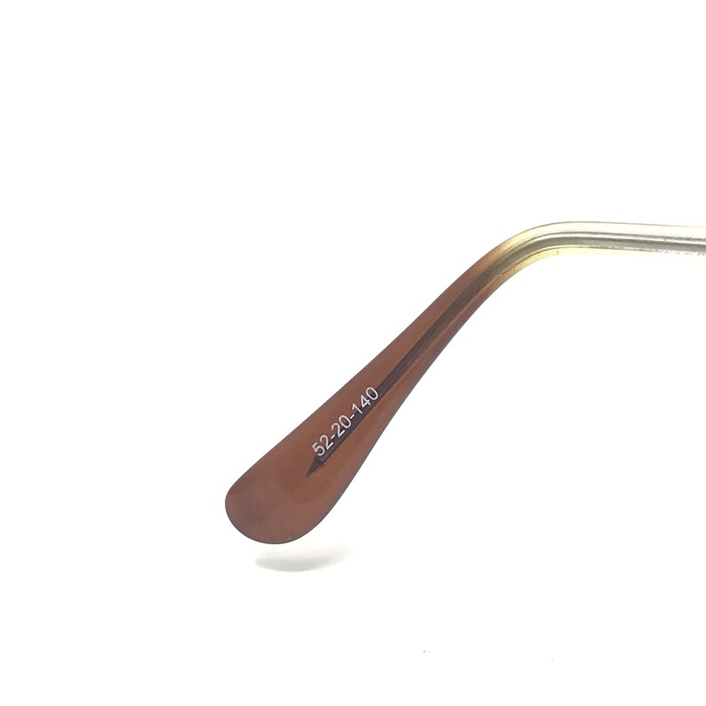 May include: Close-up of the arm of a pair of eyeglasses. The arm is a light brown color with the text "52-20-140" printed on it. The frame is a gold color.
