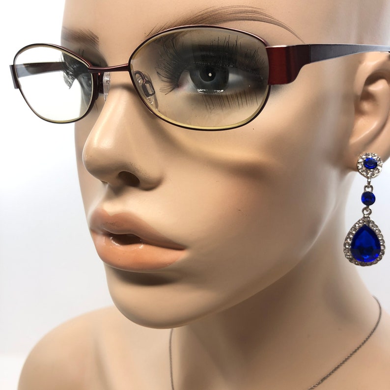 May include: A mannequin head wearing burgundy-framed eyeglasses and dangling blue rhinestone earrings. The earrings feature a small round top, a small blue gem, and a large teardrop-shaped blue gem, all surrounded by clear rhinestones. A silver necklace is also visible.