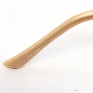 May include: Close-up of a beige-colored eyeglass arm. The text "A3L4U 135 SALENA 21663741" is printed in a dark color on the arm. The arm curves slightly and tapers to a rounded end.