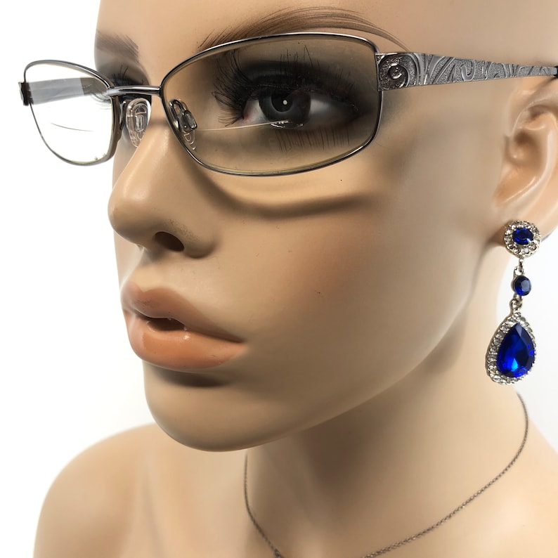 May include: Silver-framed eyeglasses with a decorative etched pattern on the temples. The image also features a pair of dangling earrings with large, vibrant blue teardrop-shaped gems, surrounded by smaller clear stones. The earrings are suspended from a smaller, round blue gem.