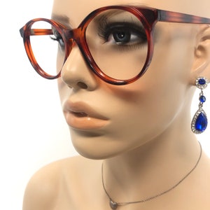 May include: Brown tortoiseshell eyeglasses with round frames. The glasses are on a mannequin head, along with a pair of blue and silver drop earrings and a silver necklace with a heart pendant.