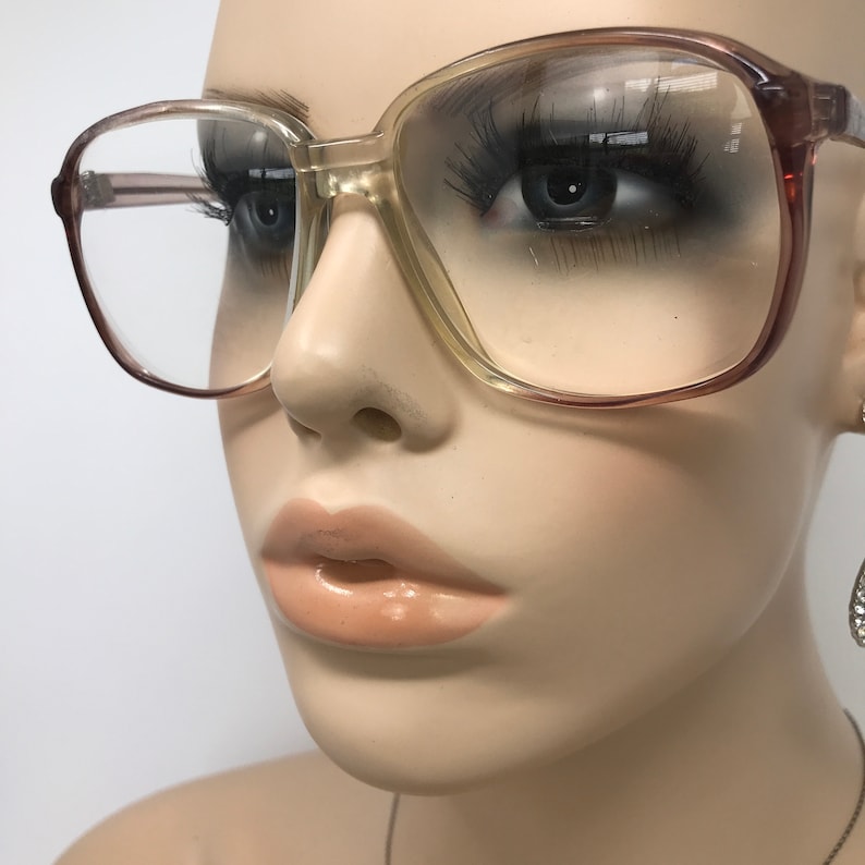 May include: Large, square-framed eyeglasses with a gradient color scheme. The frames transition from a light beige to a reddish-brown hue. The lenses are clear, and the glasses have a classic, vintage aesthetic.