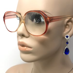 May include: A pair of large, round, peach-colored eyeglasses with clear lenses. The glasses are on a mannequin's face. The mannequin is wearing blue teardrop-shaped earrings with rhinestones.