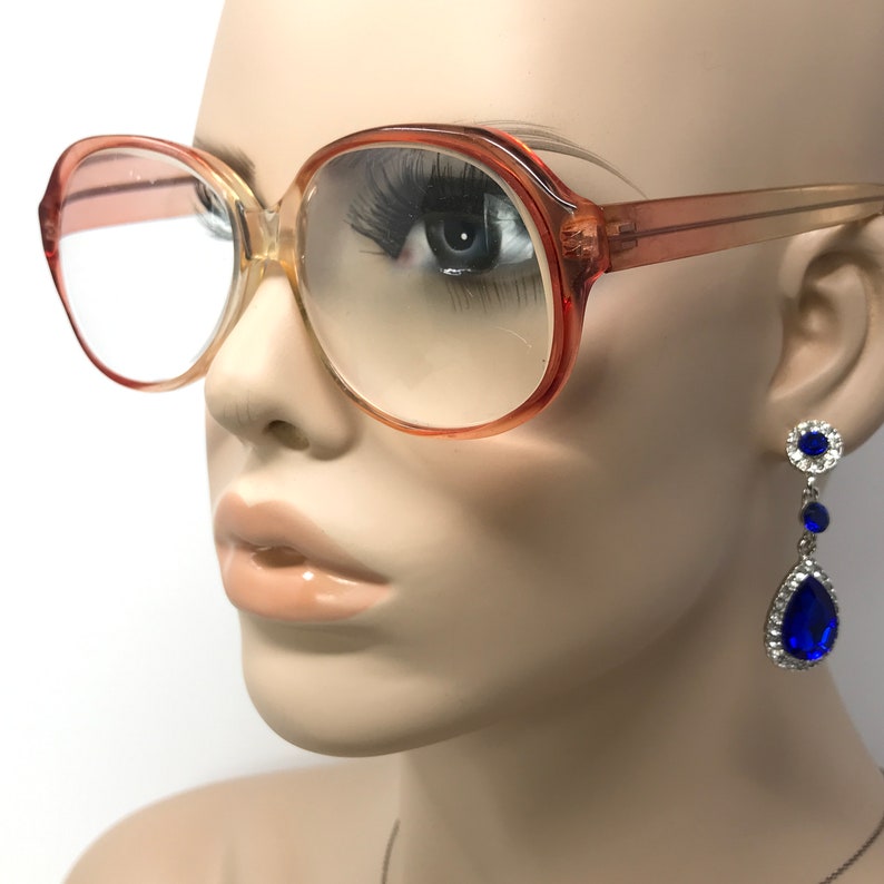 May include: A pair of large, round, peach-colored eyeglasses with clear lenses. The glasses are on a mannequin's face. The mannequin is wearing blue teardrop-shaped earrings with rhinestones.
