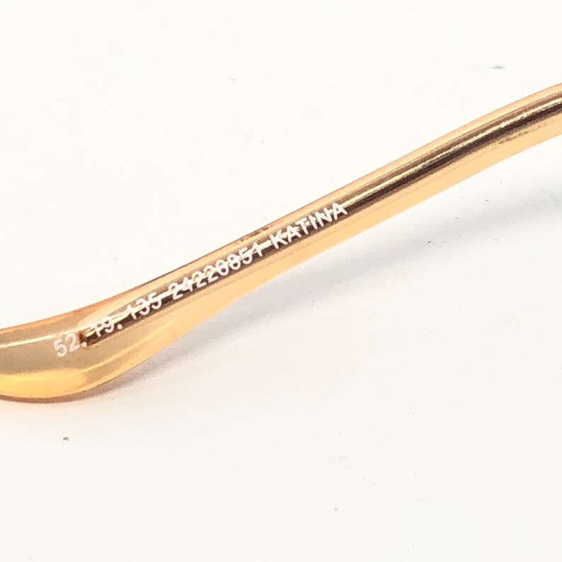 May include: Close-up of a gold-colored eyeglass frame arm. The arm has the numbers "52. 19. 135 24220054 KATINA" printed in white. The frame is on a white background.