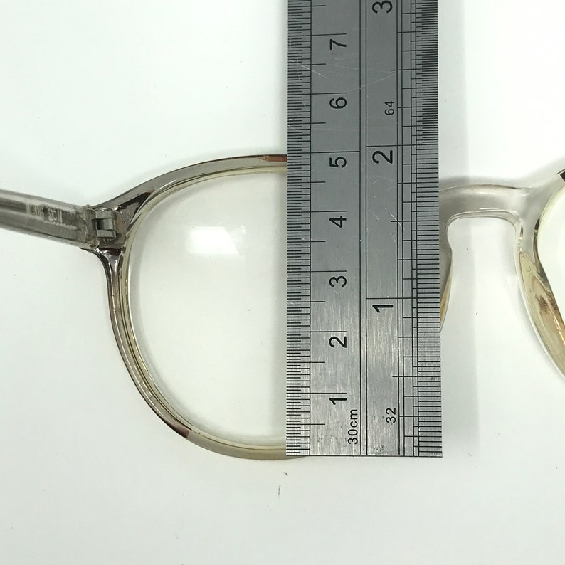 Puede incluir: Gafas transparentes con montura plateada se miden con una regla met&aacute;lica. La anchura de la lente es de aproximadamente 5 cm. Las gafas est&aacute;n sobre un fondo blanco.