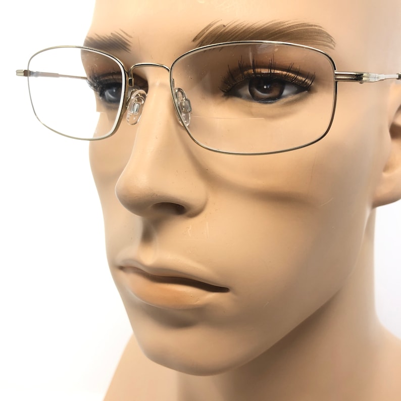 May include: Close-up of eyeglasses with a silver metal frame. The rectangular lenses are clear, and the nose pads are transparent. The glasses are on a mannequin head with a neutral skin tone. The overall style is classic and understated.
