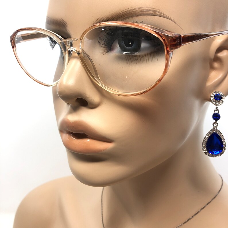 May include: A mannequin wears clear eyeglasses with a light brown frame. Long, dangling earrings with a silver setting and large, blue teardrop-shaped gems are also visible. The earrings have smaller blue and clear gems.