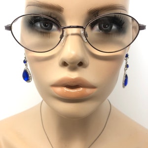 Peut inclure: Lunettes ovales avec une fine monture en m&eacute;tal fonc&eacute;. L'image pr&eacute;sente &eacute;galement une paire de boucles d'oreilles pendantes avec de grosses pierres bleues en forme de goutte et de plus petites pierres bleues. Les boucles d'oreilles sont serties dans un m&eacute;tal de couleur argent.