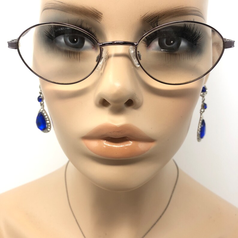 Peut inclure: Lunettes ovales avec une fine monture en m&eacute;tal fonc&eacute;. L'image pr&eacute;sente &eacute;galement une paire de boucles d'oreilles pendantes avec de grosses pierres bleues en forme de goutte et de plus petites pierres bleues. Les boucles d'oreilles sont serties dans un m&eacute;tal de couleur argent.
