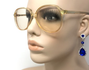 May include: A pair of large, round, amber-colored plastic framed glasses with clear lenses. The glasses are worn by a mannequin with blue teardrop earrings.