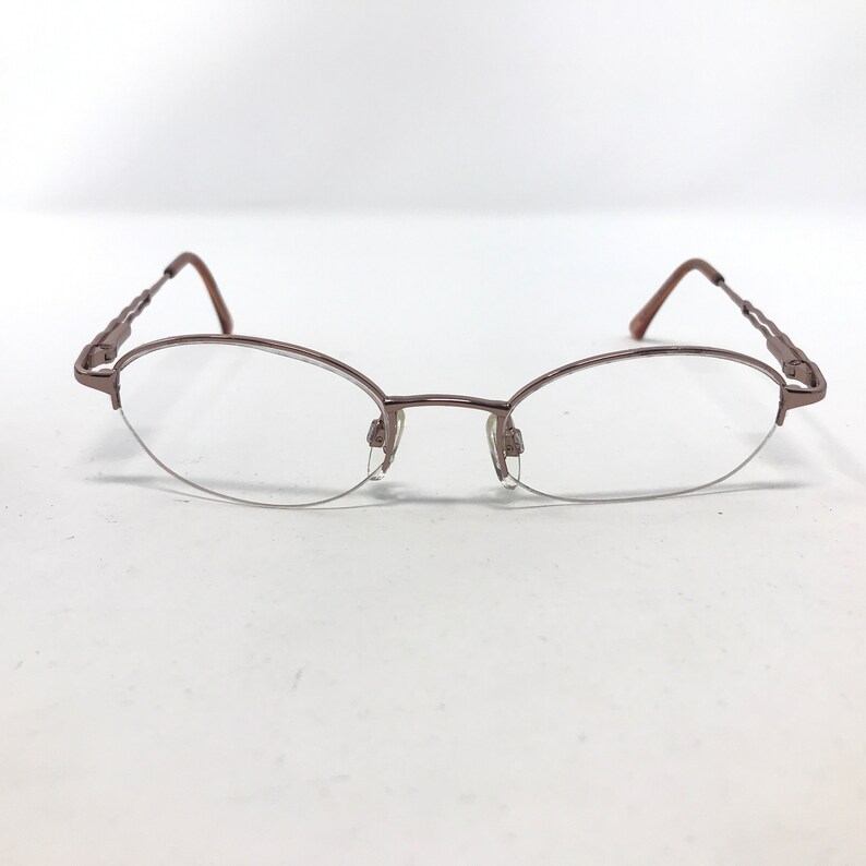 May include: A pair of oval-shaped eyeglasses with a bronze-colored metal frame. The lenses are clear, and the frame has a delicate, minimalist design. The temples feature a decorative, twisted pattern.