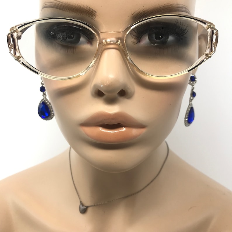 Puede incluir: Gafas transparentes con montura dorada y adornos decorativos en las patillas. El maniqu&iacute; tambi&eacute;n lleva pendientes largos azules en forma de l&aacute;grima con detalles plateados y un peque&ntilde;o collar plateado con un colgante de coraz&oacute;n.