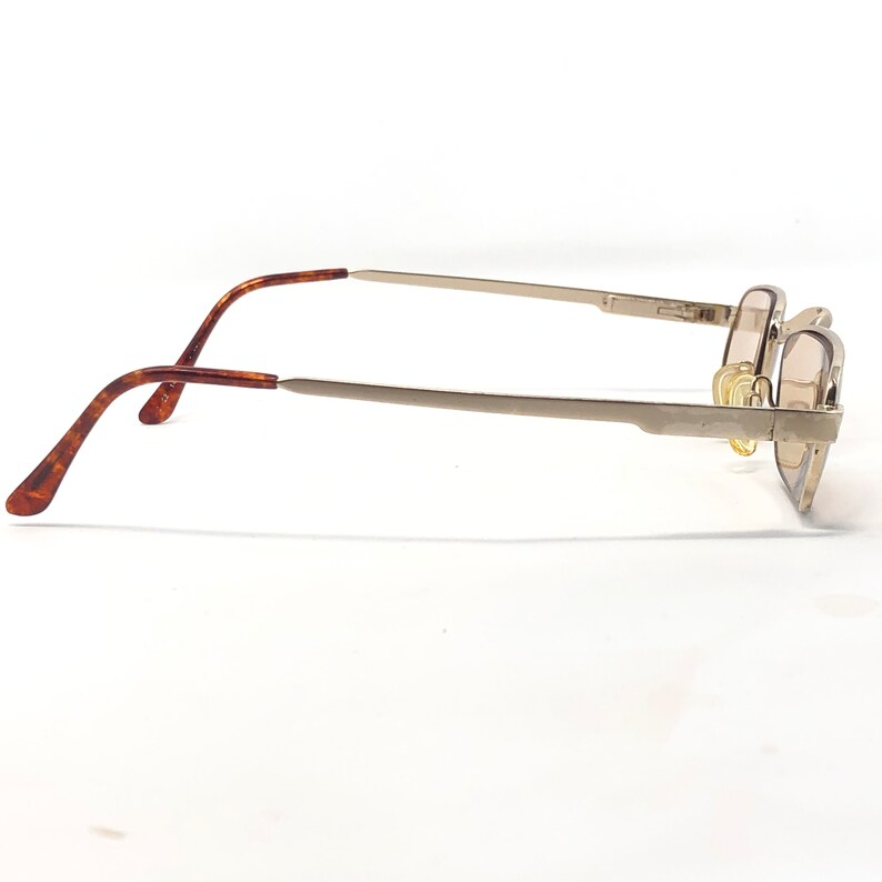 May include: Side view of eyeglasses with a silver metal frame and brown tortoiseshell temples. The lenses are a light brown tint. The glasses are set against a white background.
