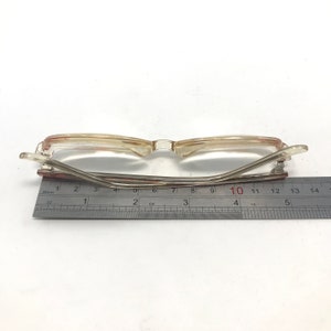 K&ouml;nnte beinhalten: Eine Brille aus klarem Kunststoff mit einem roten Streifen auf der Nasenbr&uuml;cke. Die Brille wird mit einem Lineal gemessen und zeigt eine Breite von 10 Zentimetern.
