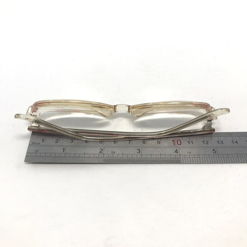 K&ouml;nnte beinhalten: Eine Brille aus klarem Kunststoff mit einem roten Streifen auf der Nasenbr&uuml;cke. Die Brille wird mit einem Lineal gemessen und zeigt eine Breite von 10 Zentimetern.