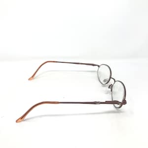 May include: A pair of round-framed eyeglasses with a brown metal frame and clear lenses. The temples are a lighter brown color. The glasses are set against a white background.