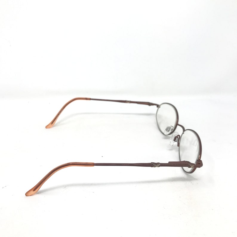 May include: A pair of round-framed eyeglasses with a brown metal frame and clear lenses. The temples are a lighter brown color. The glasses are set against a white background.
