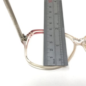 May include: Clear eyeglasses with a red accent on the frame, measured with a ruler. The frame is approximately 2 inches wide. The glasses are set against a white background.