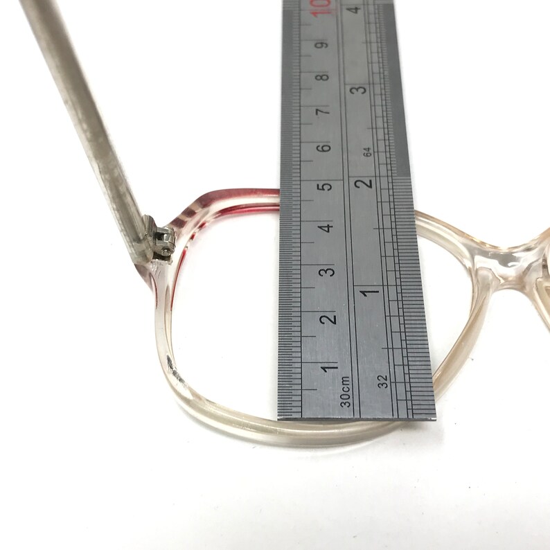 May include: Clear eyeglasses with a red accent on the frame, measured with a ruler. The frame is approximately 2 inches wide. The glasses are set against a white background.