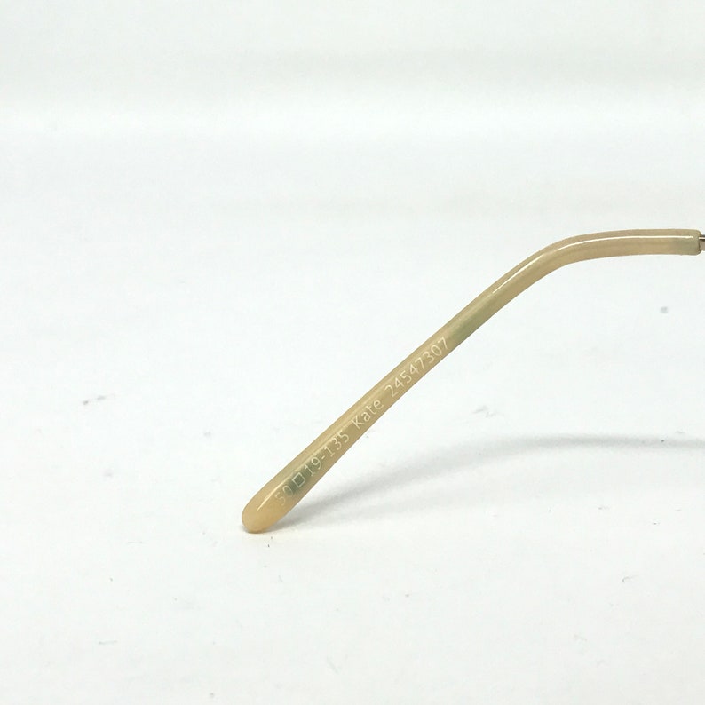 May include: A close-up of the arm of a pair of eyeglasses. The arm is a light beige color and has the text "50▢19-135 Kate 24547307" printed on it. The end of the arm is rounded.
