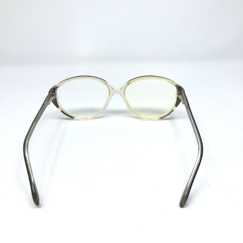 May include: Clear eyeglasses with a light-colored frame and thin, silver-toned arms. The lenses are oval-shaped and the frame has a subtle, patterned detail near the hinges. The glasses are set against a white background.