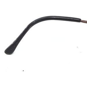 Puede incluir: Primer plano de una patilla de gafas negra con un dise&ntilde;o curvo. La patilla tiene una punta redondeada y muestra signos de desgaste. La montura parece estar hecha de un material oscuro, posiblemente pl&aacute;stico. El fondo es blanco liso.