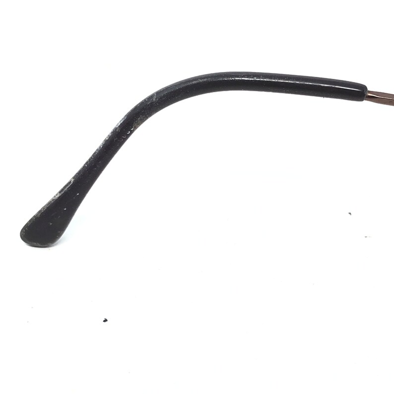 Puede incluir: Primer plano de una patilla de gafas negra con un dise&ntilde;o curvo. La patilla tiene una punta redondeada y muestra signos de desgaste. La montura parece estar hecha de un material oscuro, posiblemente pl&aacute;stico. El fondo es blanco liso.