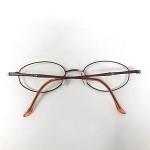 May include: Oval-shaped eyeglasses with a brown metal frame and clear lenses. The temples are a lighter brown color. The glasses are designed with a minimalist aesthetic.
