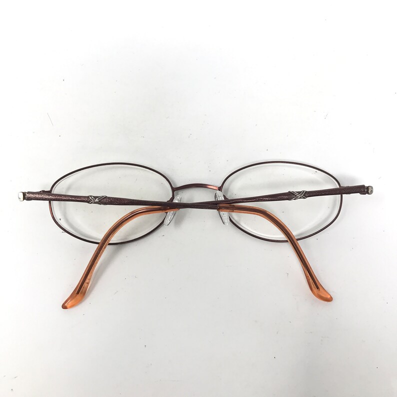 May include: Oval-shaped eyeglasses with a brown metal frame and clear lenses. The temples are a lighter brown color. The glasses are designed with a minimalist aesthetic.