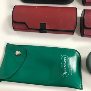 May include: An assortment of eyeglass cases in various colors. A red rectangular case with a black border and a black clasp is visible. A green, soft case with the text "Specsavers" is also present.