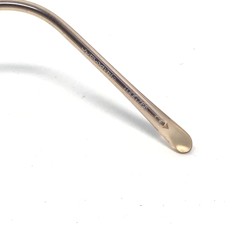 May include: Close-up of a gold-toned metal eyeglass arm. The arm has text printed on it, including the word "CHAMPAGNE" and the number "39/16". The end of the arm is rounded and has a light gold color.