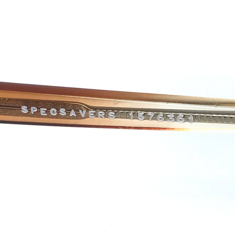May include: Close-up of the inside of a gold-colored pair of glasses with the text "SPECSAVERS 11576361" printed on the inside of the arm.