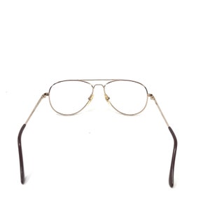 May include: Silver-framed aviator eyeglasses with clear lenses. The glasses have a double bridge and dark brown earpieces. The lenses are oval-shaped and the frame is a shiny silver color. The glasses are set against a white background.